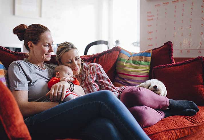 Same sex couple enjoying a cuddle together at home, with their baby daughter and their pet dog.